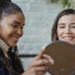 happy young female client looking in mirror and smiling near positive ethnic makeup artist