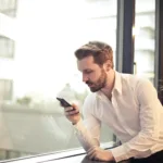 photo of man in white dress shirt holding phone near window